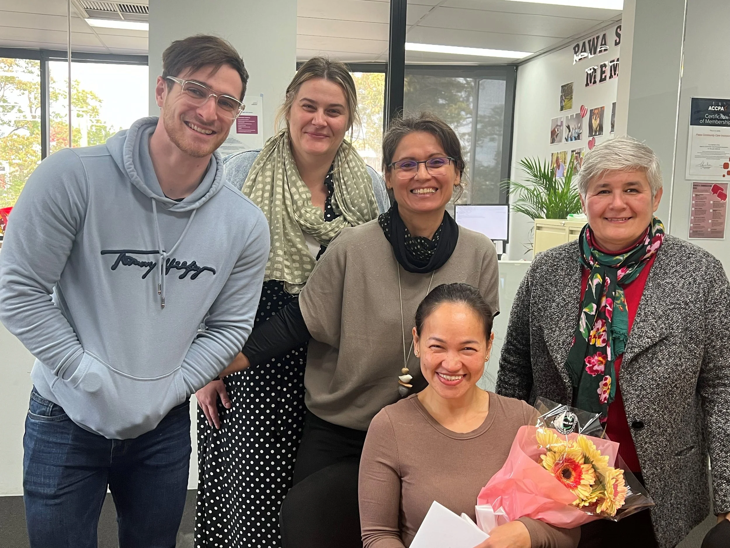 Group of five women and one man in an office, with one woman holding a bouquet of flowers.