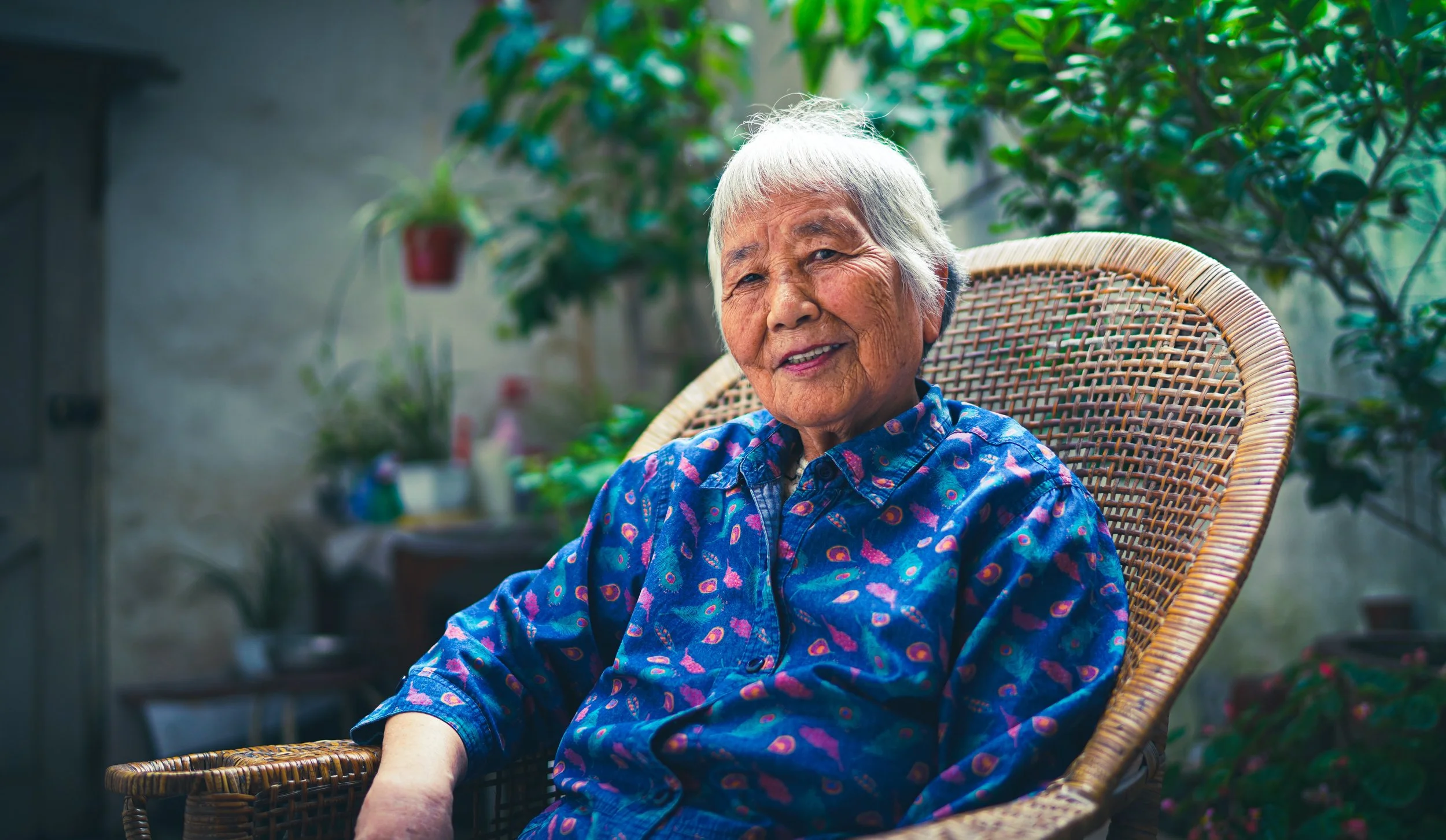 An elderly woman with white hair sitting comfortably outdoors on a wicker chair, smiling, surrounded by green plants and potted flowers.