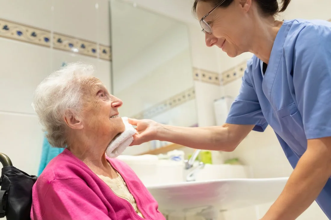 Nurse or caregiver gently wiping the face of an elderly woman in a pink sweater in a healthcare setting.