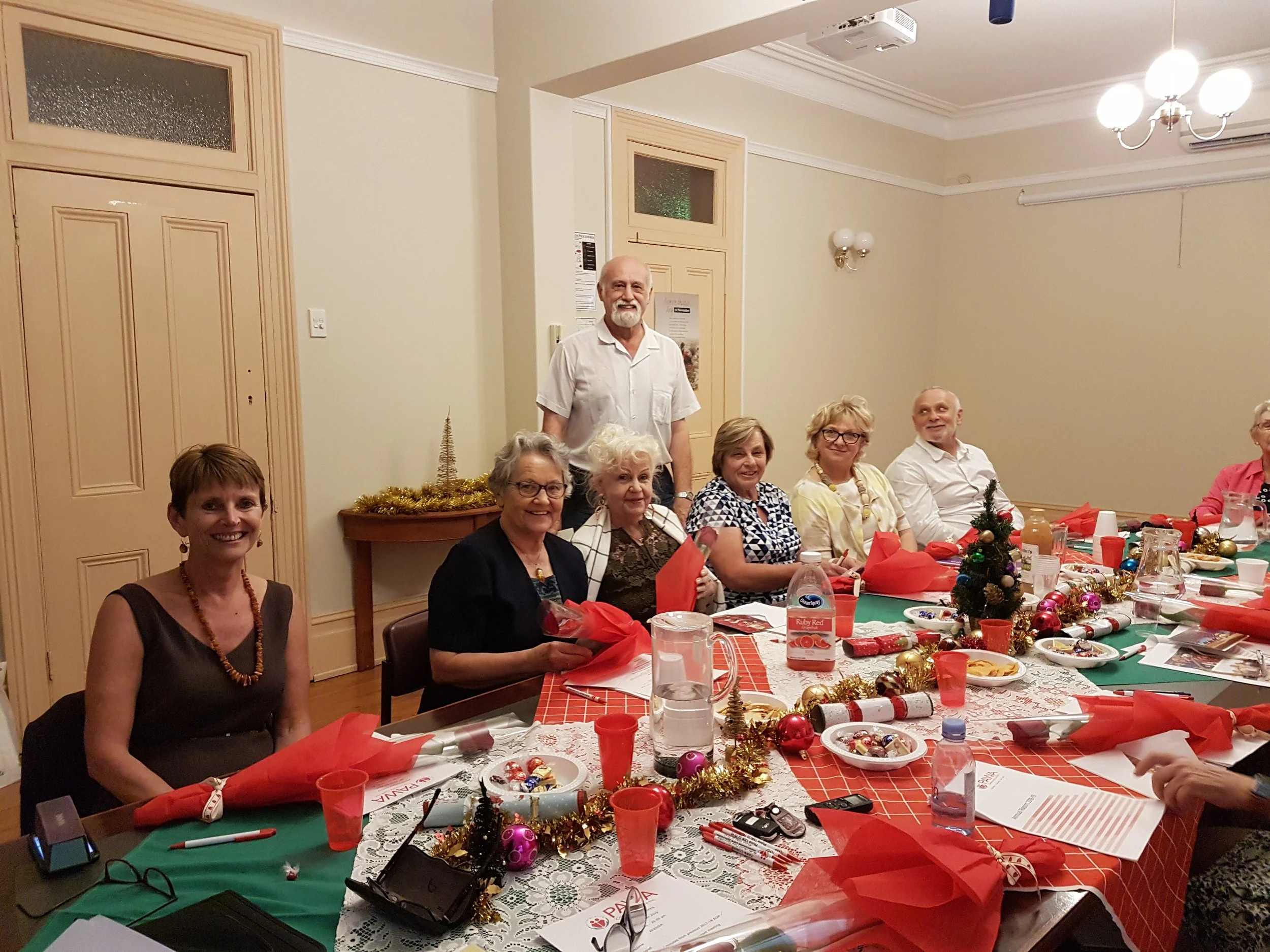 A group of elderly and middle-aged people gathered around a decorated Christmas table for a celebration. The table is covered with a red and white tablecloth and decorated with a small Christmas tree, ornaments, tinsel, and wrapped gifts. Participants are smiling and engaging with each other in a warmly lit room with cream-colored walls, wooden doors, and festive decorations.