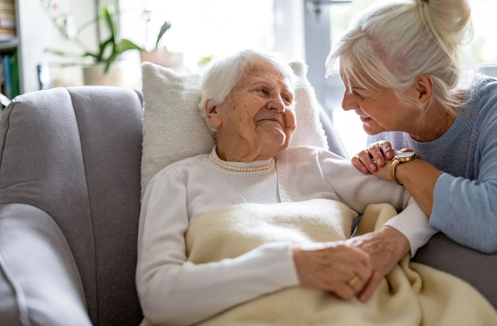 An elderly woman with white hair lying in bed, smiling and holding hands with a woman leaning over her who is smiling and touching her shoulder. The room is bright with natural light and has potted plants in the background.