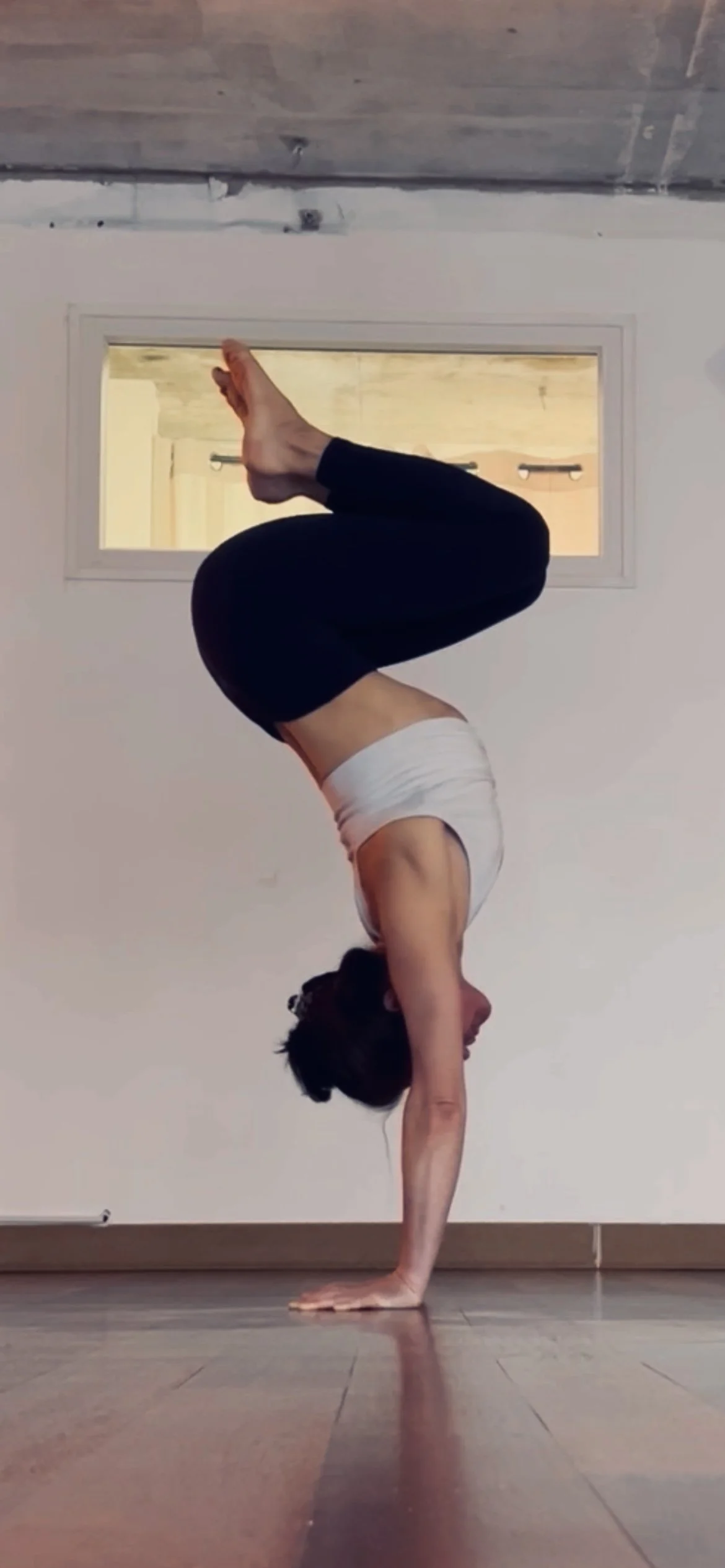 Femme faisant une posture de yoga en équilibre sur une main, formant un pont avec ses jambes repliées. Elle est dans une pièce avec un sol en bois et un mur blanc, et une fenêtre derrière elle.