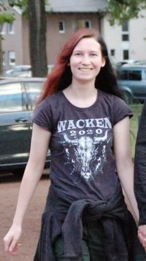 Marie schaut lächelnd nach vorn, trägt ein schwarzes T-Shirt mit dem Text "WACKEN 2020" und befindet sich im Freien vor Häusern und parkenden Autos. Ihre langen Haare sind zweifarbig, die rechte Seite rot.