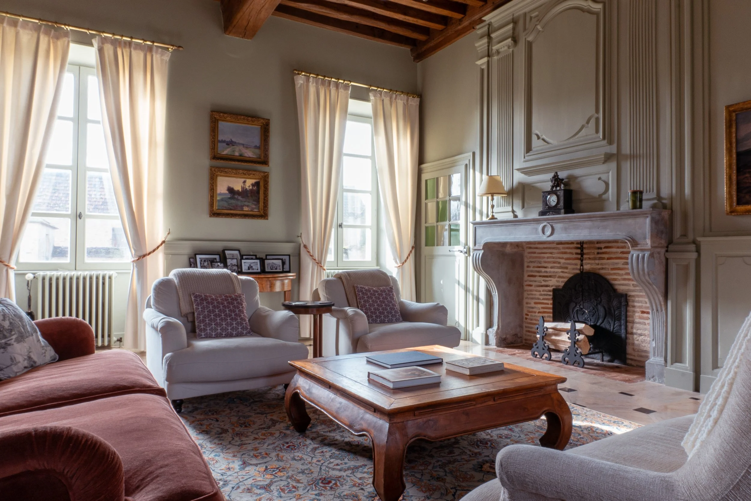 A cozy living room with "French Grey" by Farrow and Ball walls, white curtains, and a stone fireplace. There are multiple armchairs, a wooden coffee table, and framed pictures on the walls and mantle.