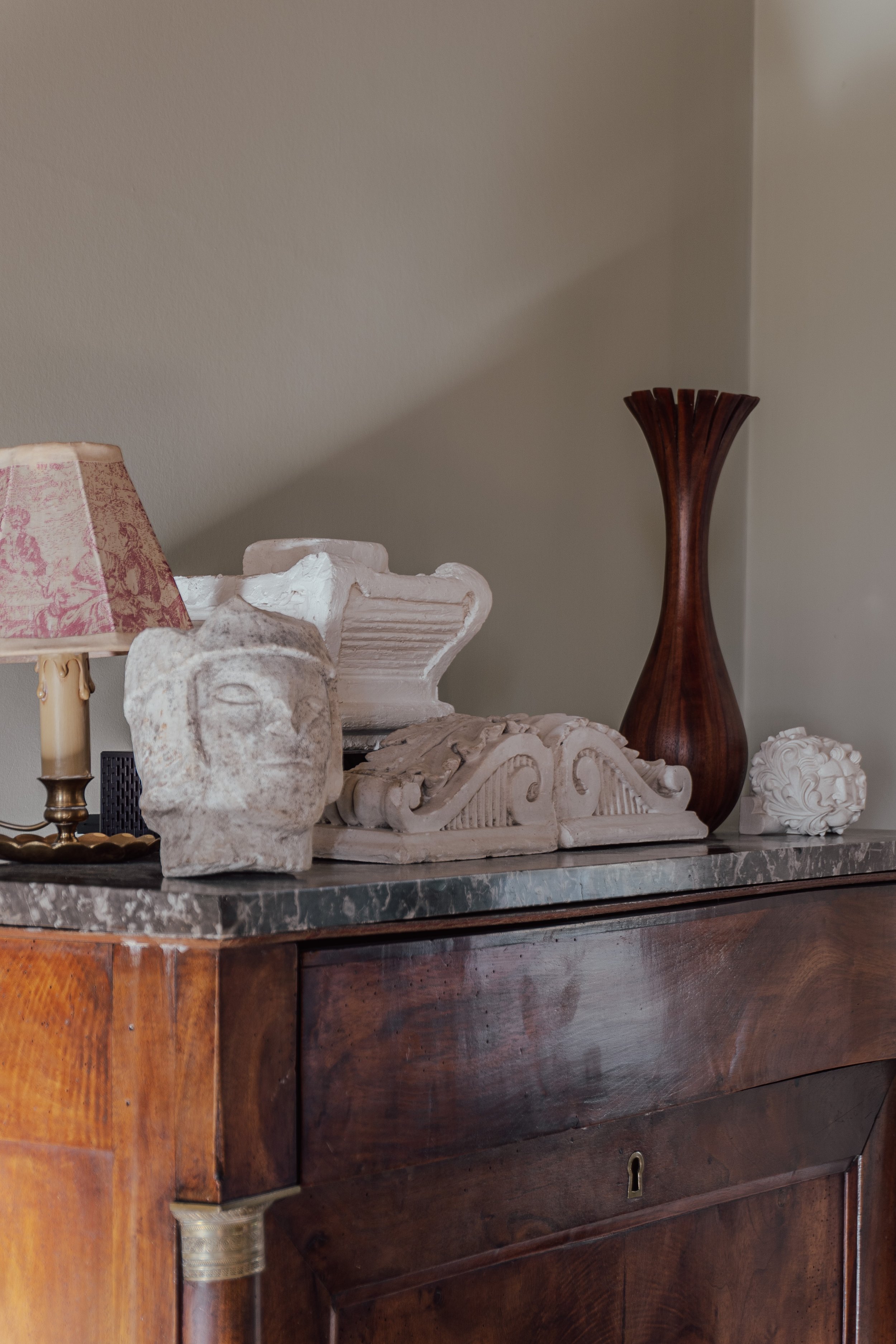 Decorative objects on top of a wooden sideboard, including a stone bust, a white architectural fragment, a dark wooden vase, a white carved sculpture, and a small floral sculpture, with a lamp on the left.