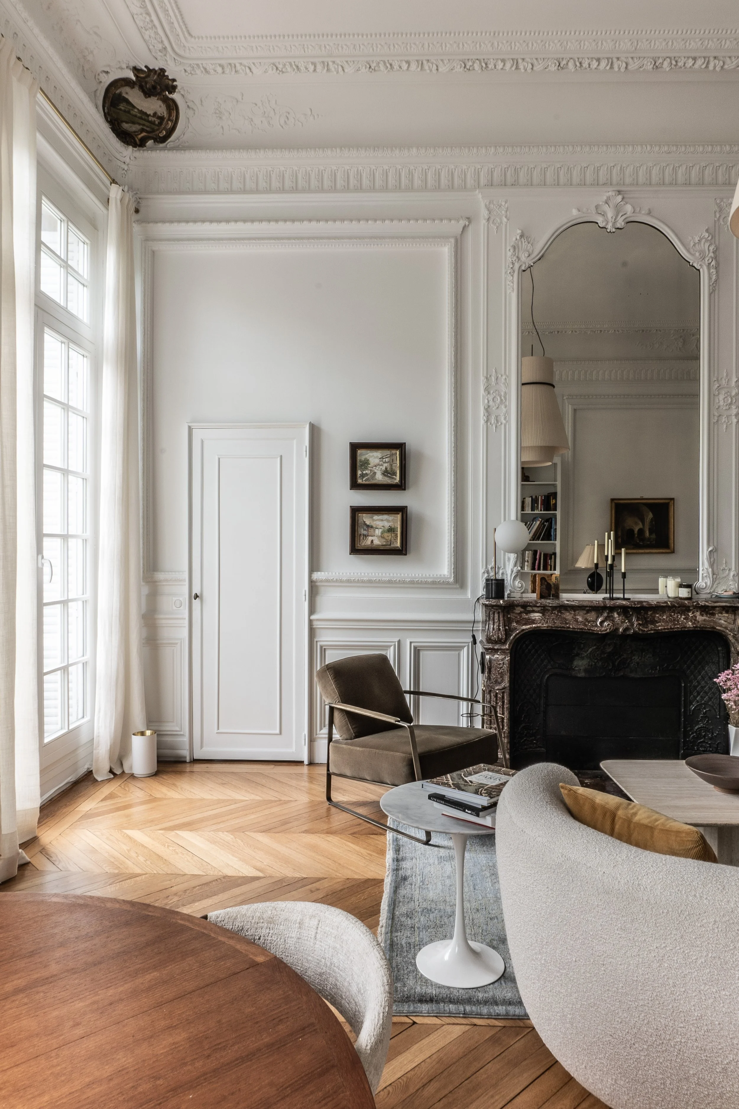 Elegant living room with white ornate walls, large window with curtains, wooden floor, green armchair, fireplace with mirror and candles, small side table with books, and partial view of a wooden dining table with chairs.
