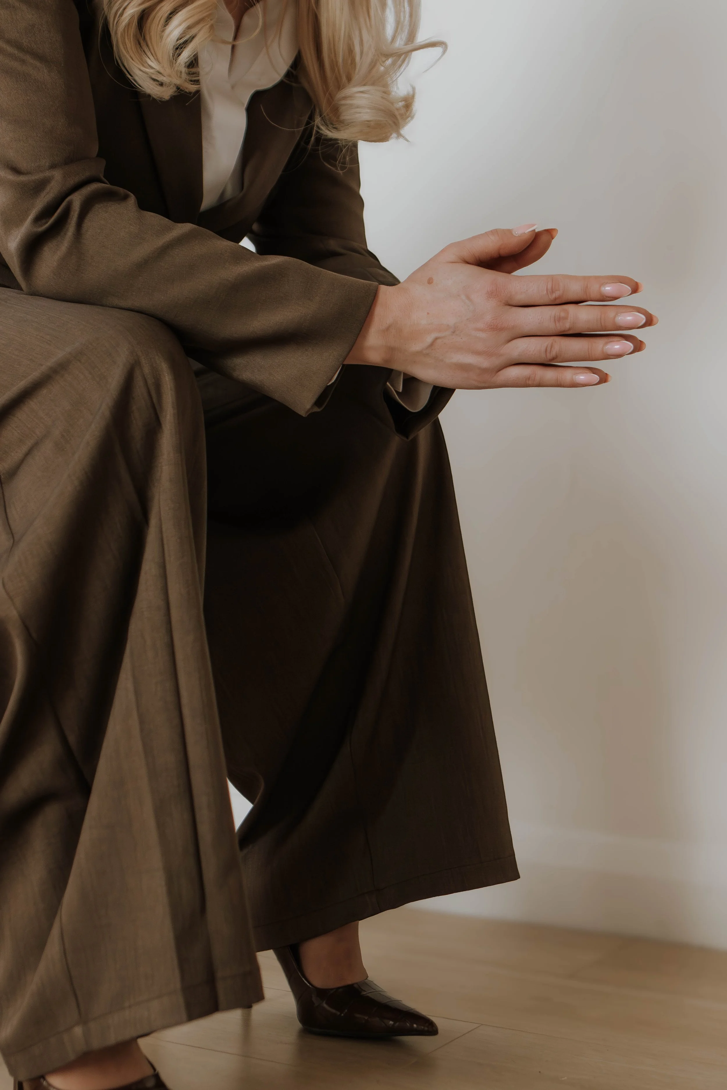 A woman in a brown suit with blonde hair is sitting on the floor, wearing brown heeled shoes, with a white wall in the background.