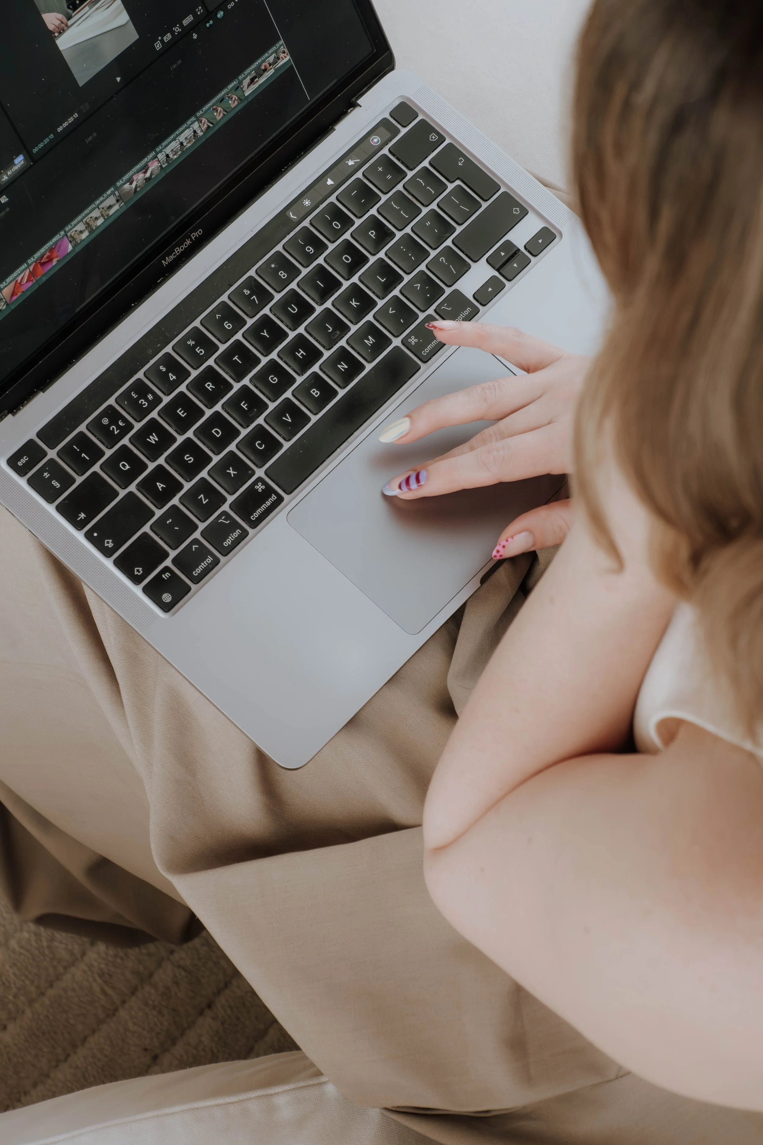Close-up of a person using a MacBook Pro laptop, with focus on their hand typing on the keyboard. The person has painted nails with a patriotic theme, and the laptop is placed on their lap, wearing beige pants. The screen displays a video editing software.