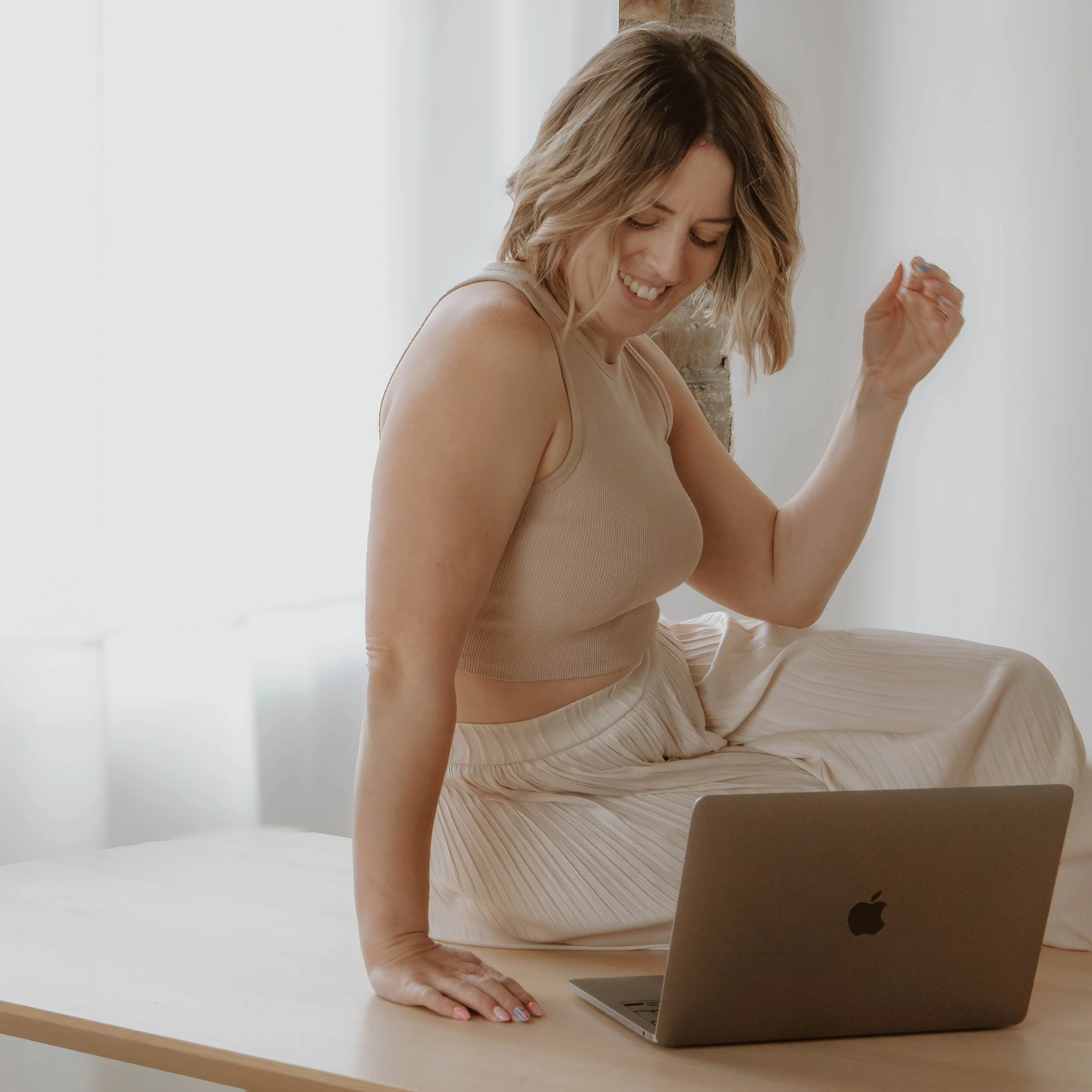 A woman with shoulder-length blonde hair looking down and smiling while working on a laptop at a light-colored desk in a bright room.