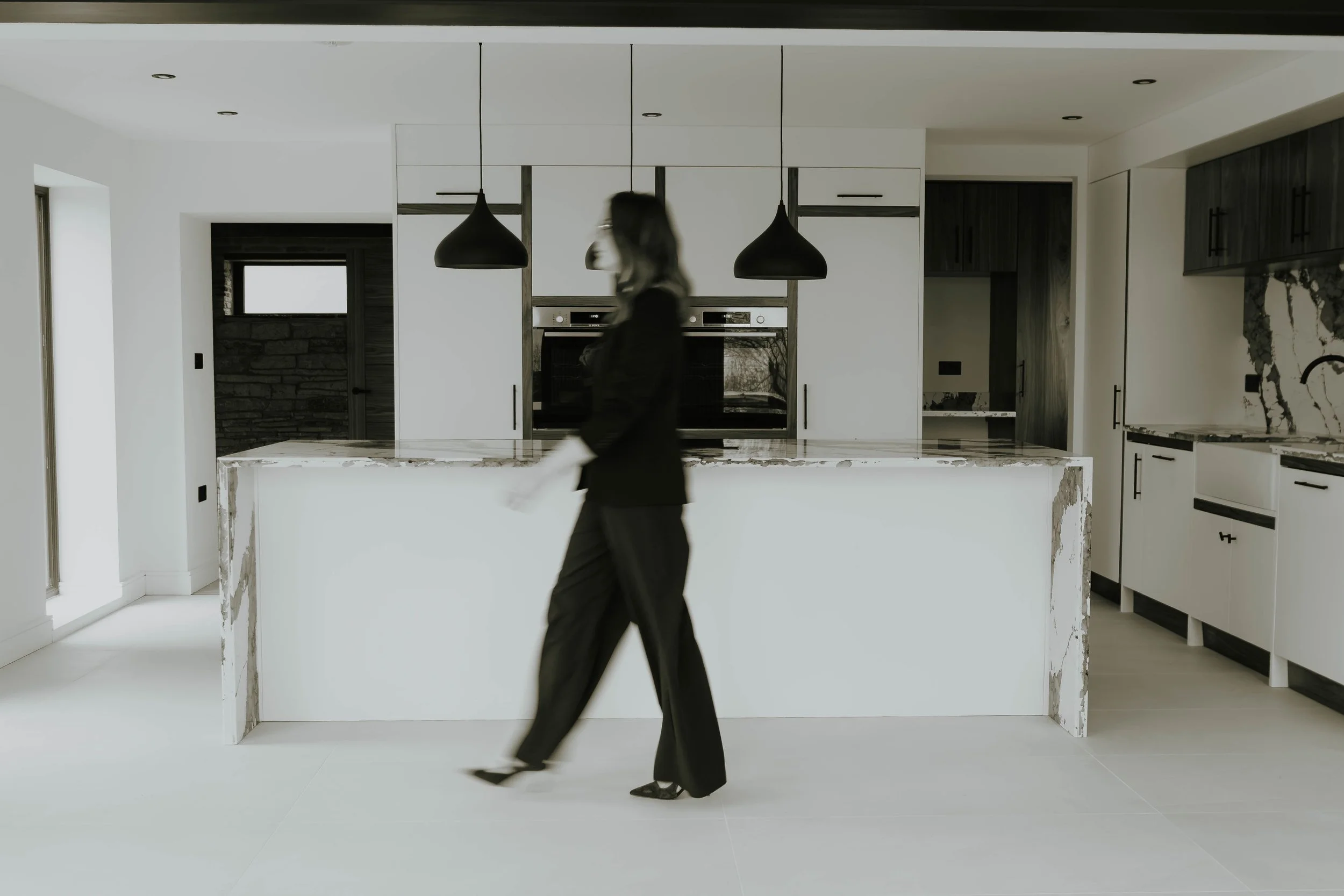 Modern kitchen with white cabinetry, black pendant lights, and a marble island counter. A woman in black walking past.