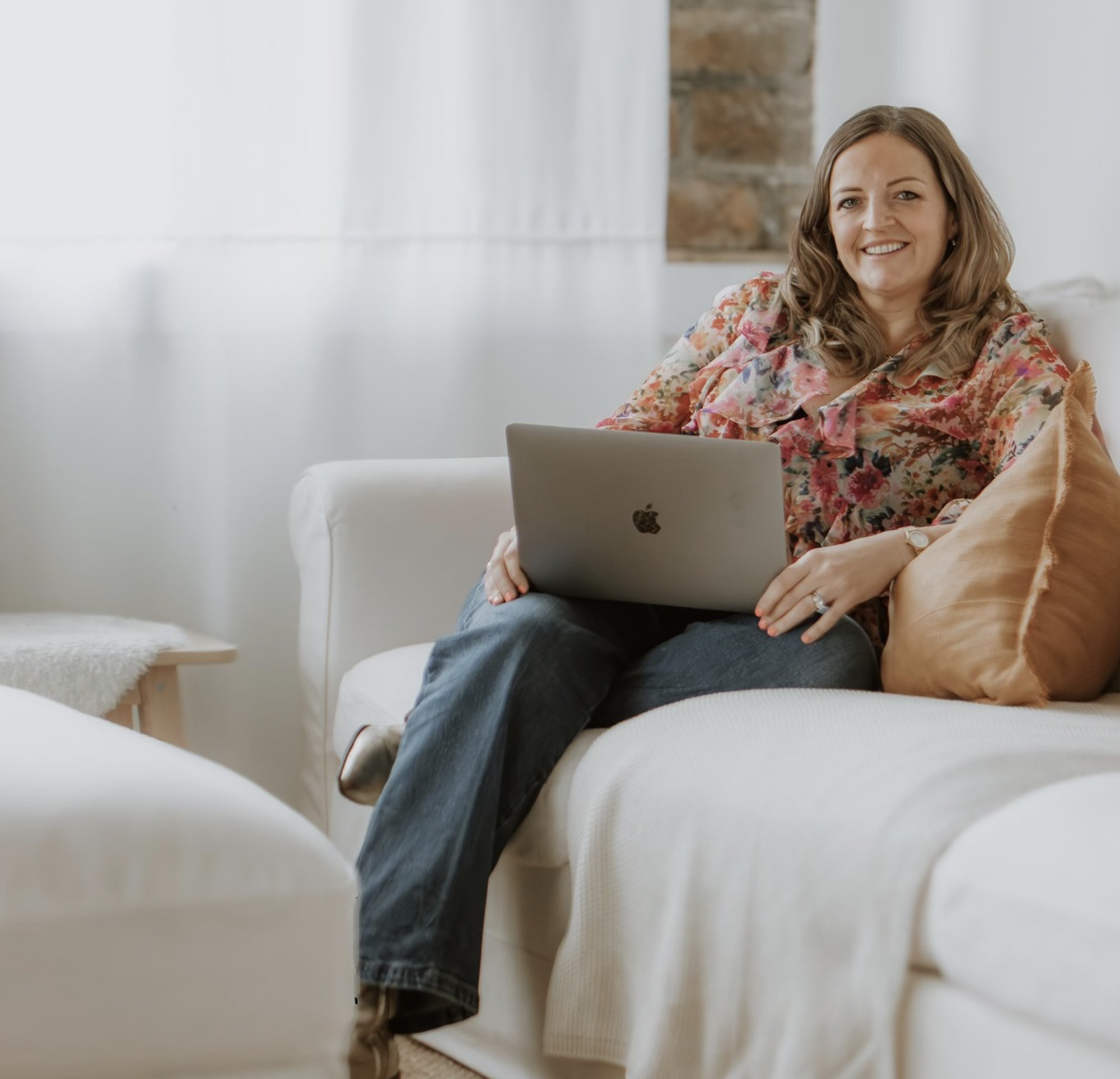 A woman with wavy brown hair smiling, wearing a floral blouse, sitting on a white couch with her legs crossed, using a silver MacBook laptop in a bright living room with a brick wall in the background.