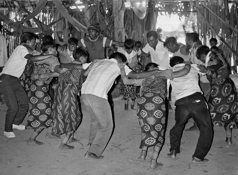 A group of people, including men, women, and children, are holding hands and dancing in a circle inside a tent or sheltered area with hanging decorations.