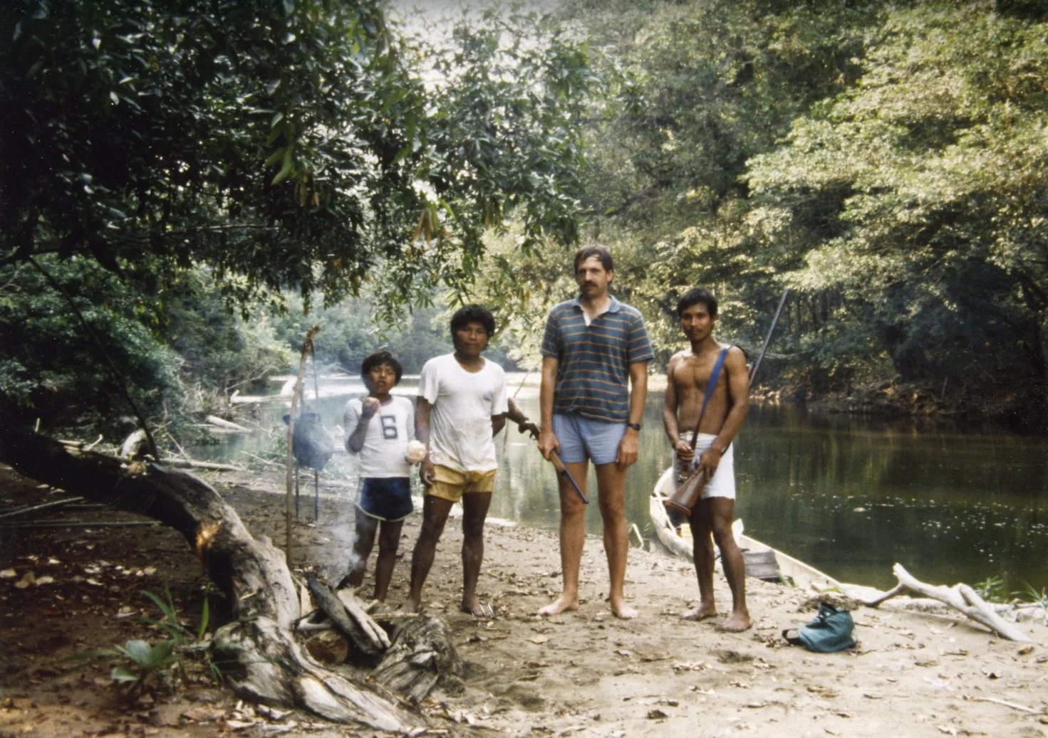 Four men standing by a river in a forested area. One man is shirtless holding a rifle, another is wearing a striped polo shirt, and two children are standing to the left. There is a small boat in the water, and the scene appears to be outdoors in a natural setting.