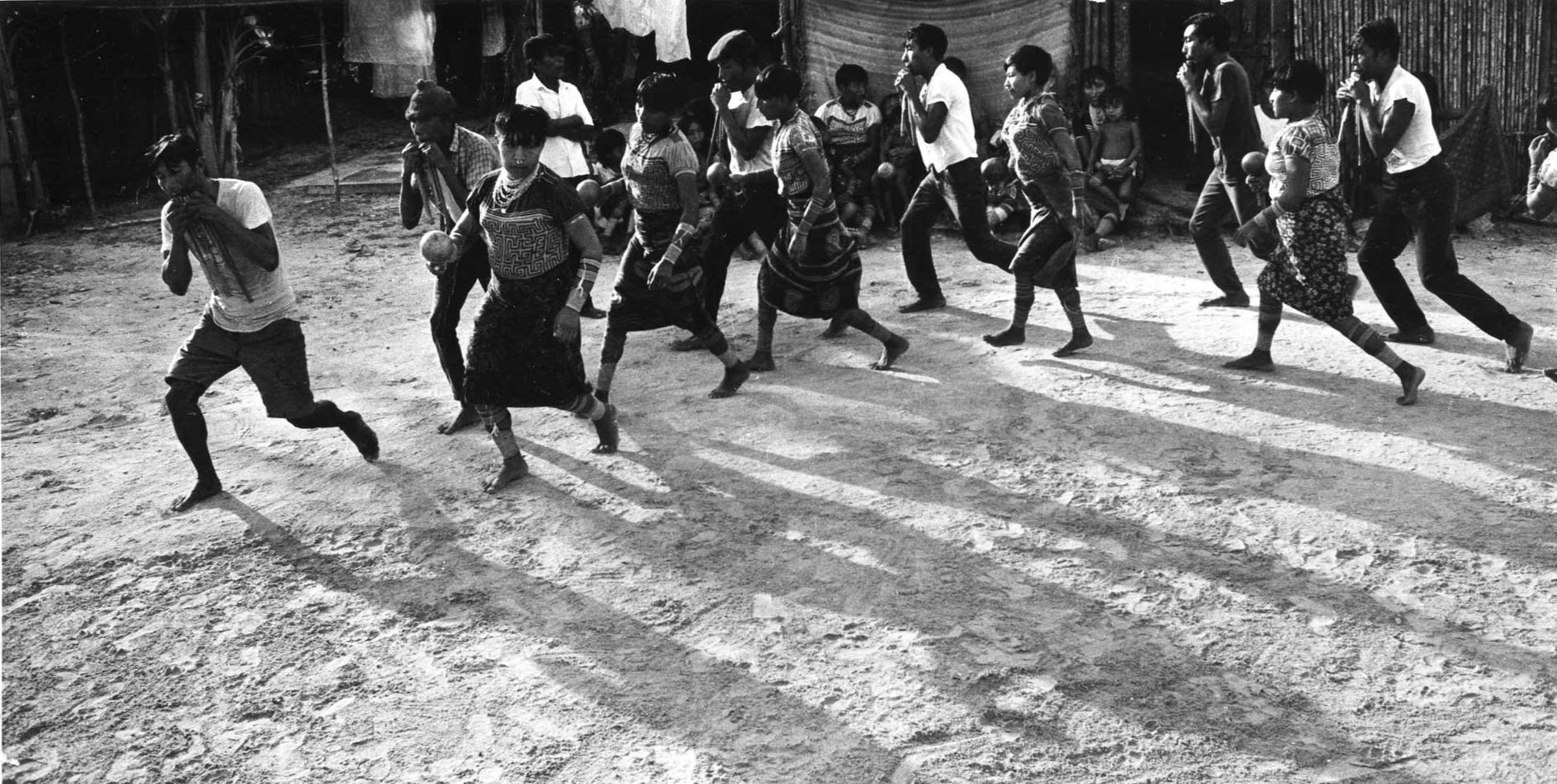 A group of children and teenagers running on a dirt ground, some holding balls, in a rural setting with simple structures in the background.