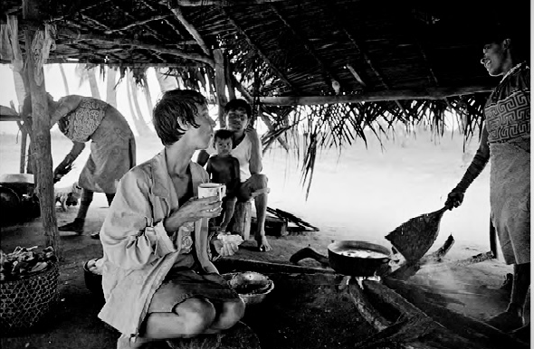 Group of people inside a thatched hut by a body of water, engaging in cooking and eating activities.