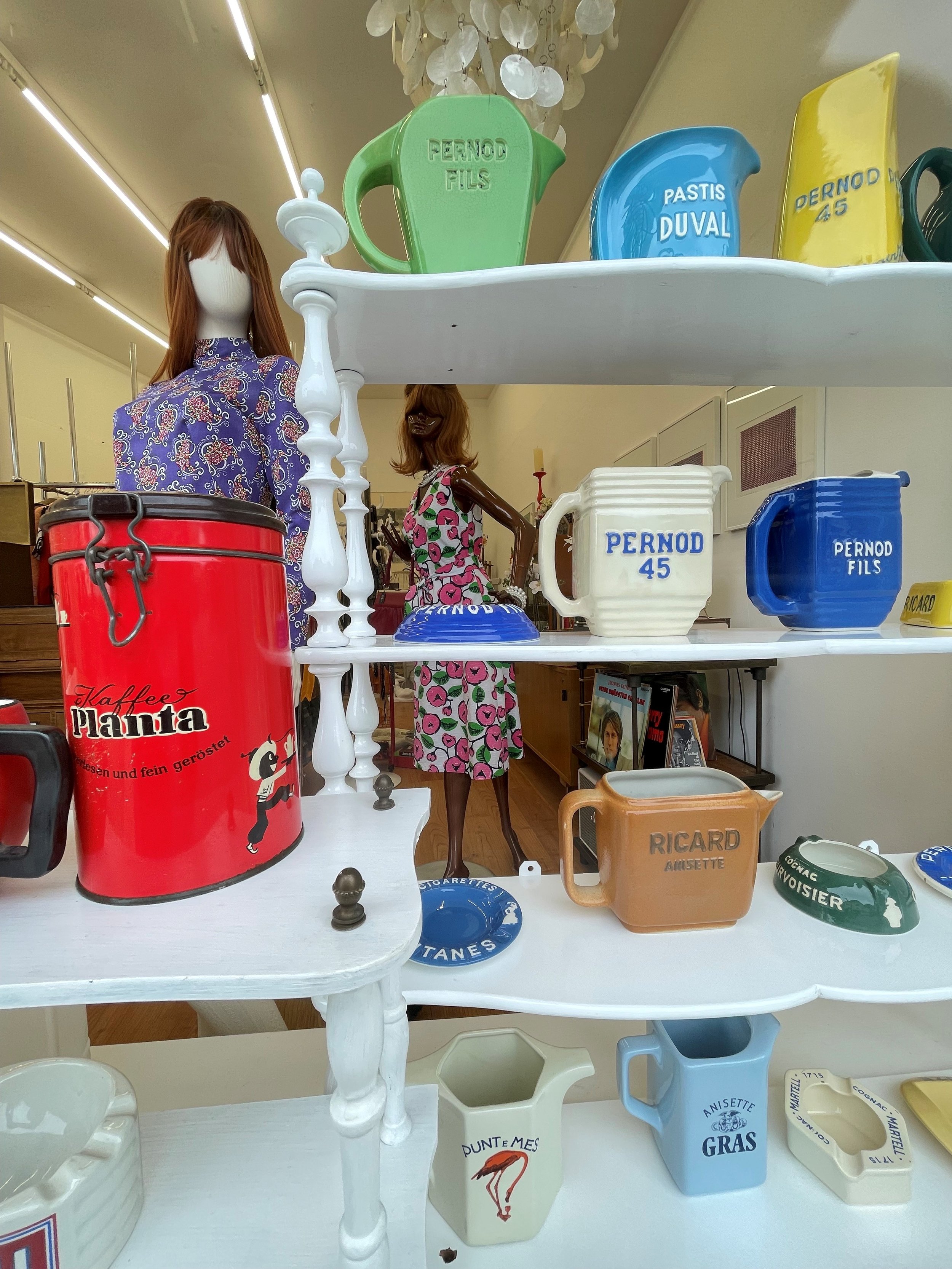 Verschiedene bunte Tassen und Becher auf einem weißen Regal, darunter eine rote Dose, ein blauer Becher, eine weiße Tasse mit Flamingo-Aufdruck, und eine braune Tasse mit Aufschrift "Ricard".