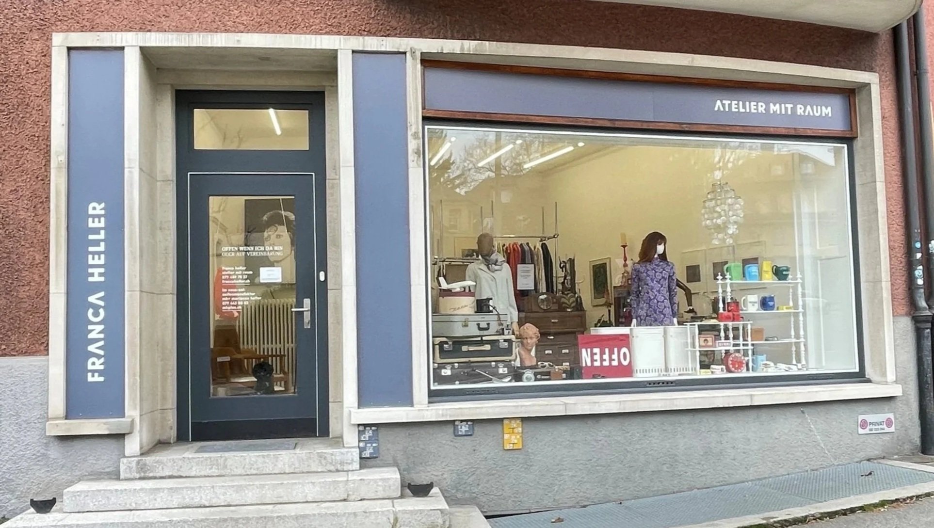 Schaufenster eines Vintage-Ladens mit Kleidung, Möbeln und Dekorationsgegenständen, im Inneren stehen zwei Personen, eine trägt eine Maske.