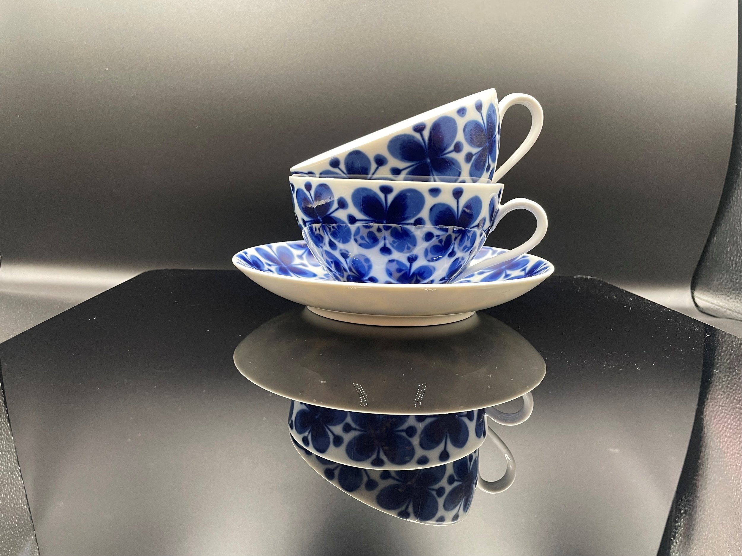 Drei blaue und weiße Teetassen mit Blumenmuster, auf einem weißen Teller, reflektiert auf einer schwarzen Oberfläche.