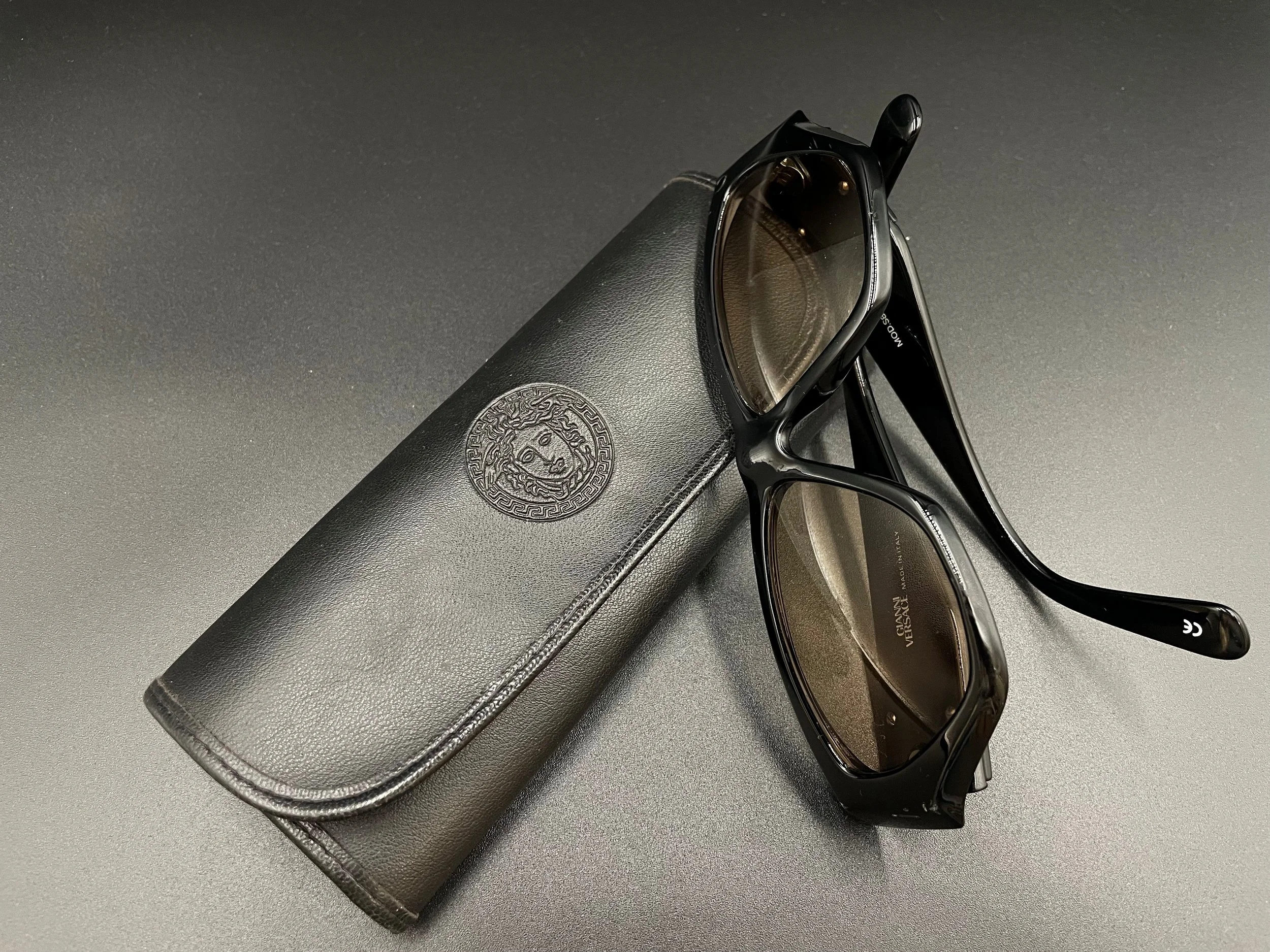 Eine schwarze Brille, ein schwarzes Etui mit einem Löwen-Logo, eine Sonnenbrille, alles auf einer schwarzen Oberfläche.