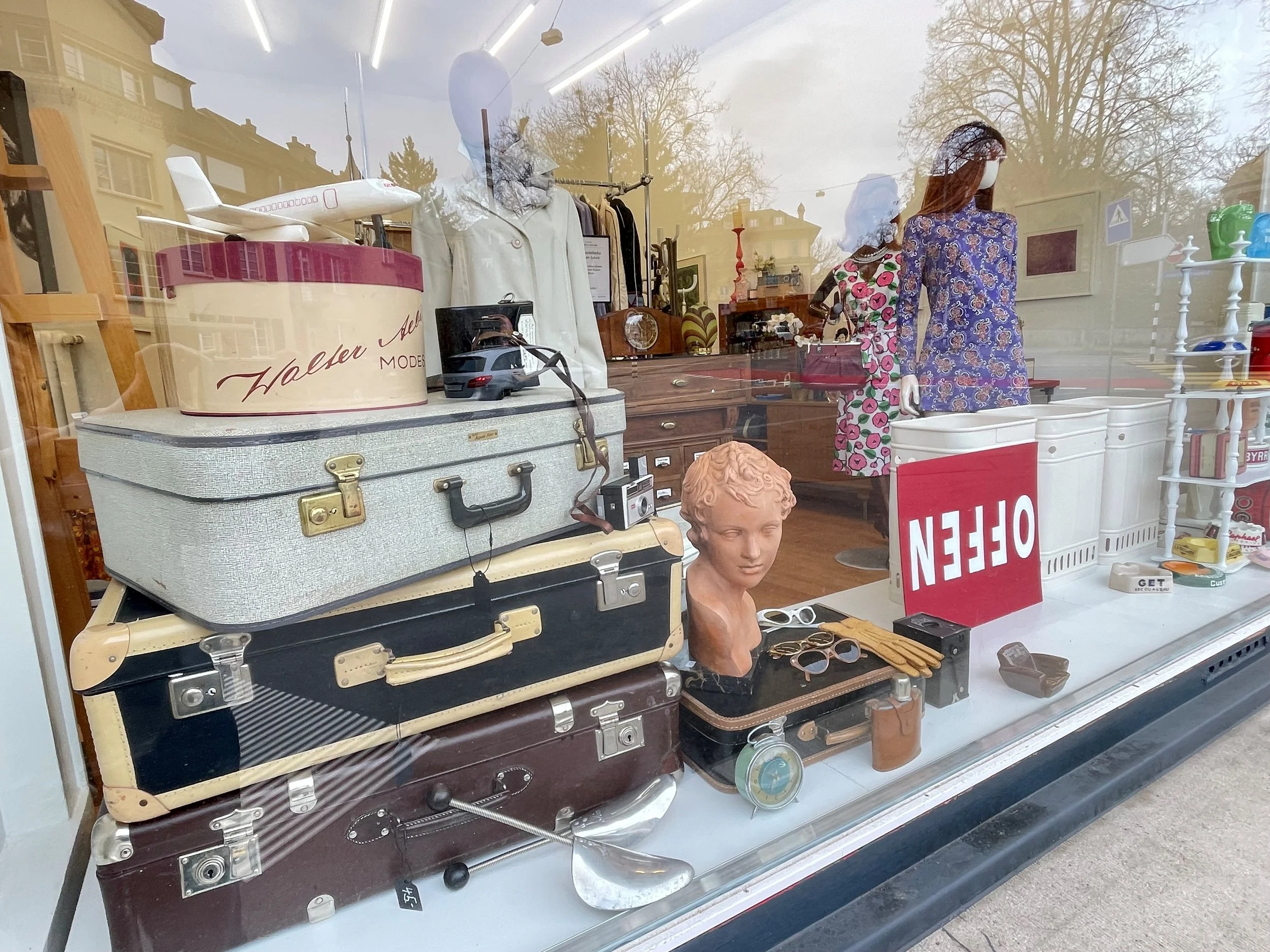 Schaufenster eines Second-Hand-Geschäfts mit verschiedenen alten Gegenständen, darunter Puppen, Taschen, Skulpturen, Brillen, Uhren, und andere Vintage-Artikel.