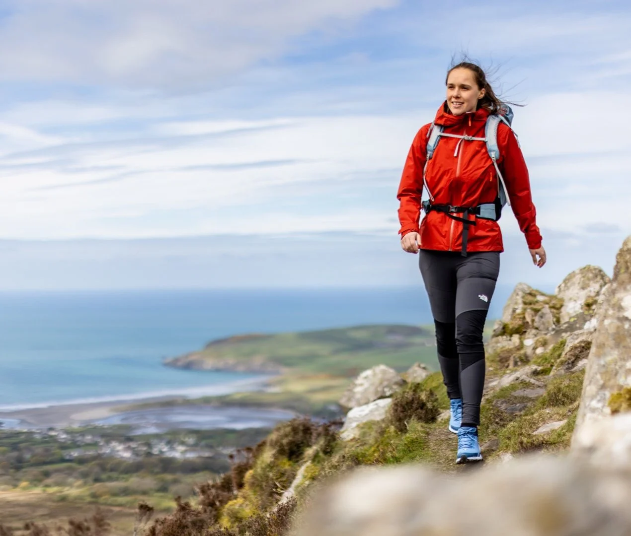 Emma Cole walking along a coastal ridge path in a red jacket, with the sea and a bay below