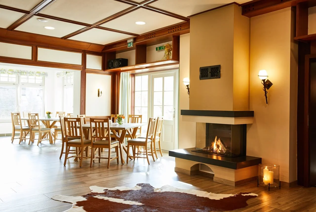 Ein heller Raum mit mehreren Holztischen und Stühlen, einem Kamin mit offenem Feuer, Kerzen, Wandlampen, großen Fenstern und Holzbalkendecke.