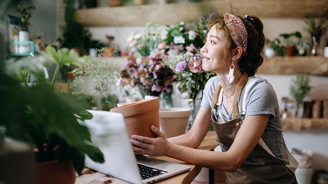 young asian plant shop owner in apron seated at computer holding potted plant surrounded by plants and flowers