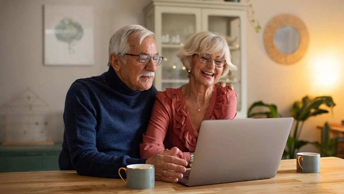 attractive older couple seated at table in warm home using laptop computer and drinking coffee