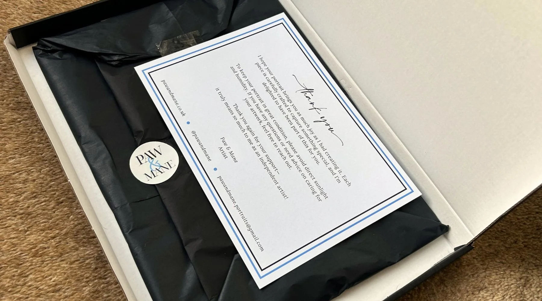 Open gift box with black tissue paper and a handwritten note thanking the recipient, accompanied by a round sticker with the words 'PAW & MATE' on it.