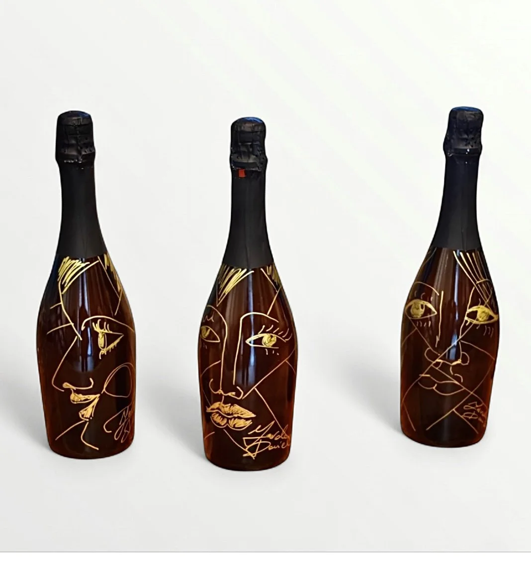 Tre bottiglie di champagne decorate con disegni di volti femminili in stile line art, posizionate su uno sfondo bianco.