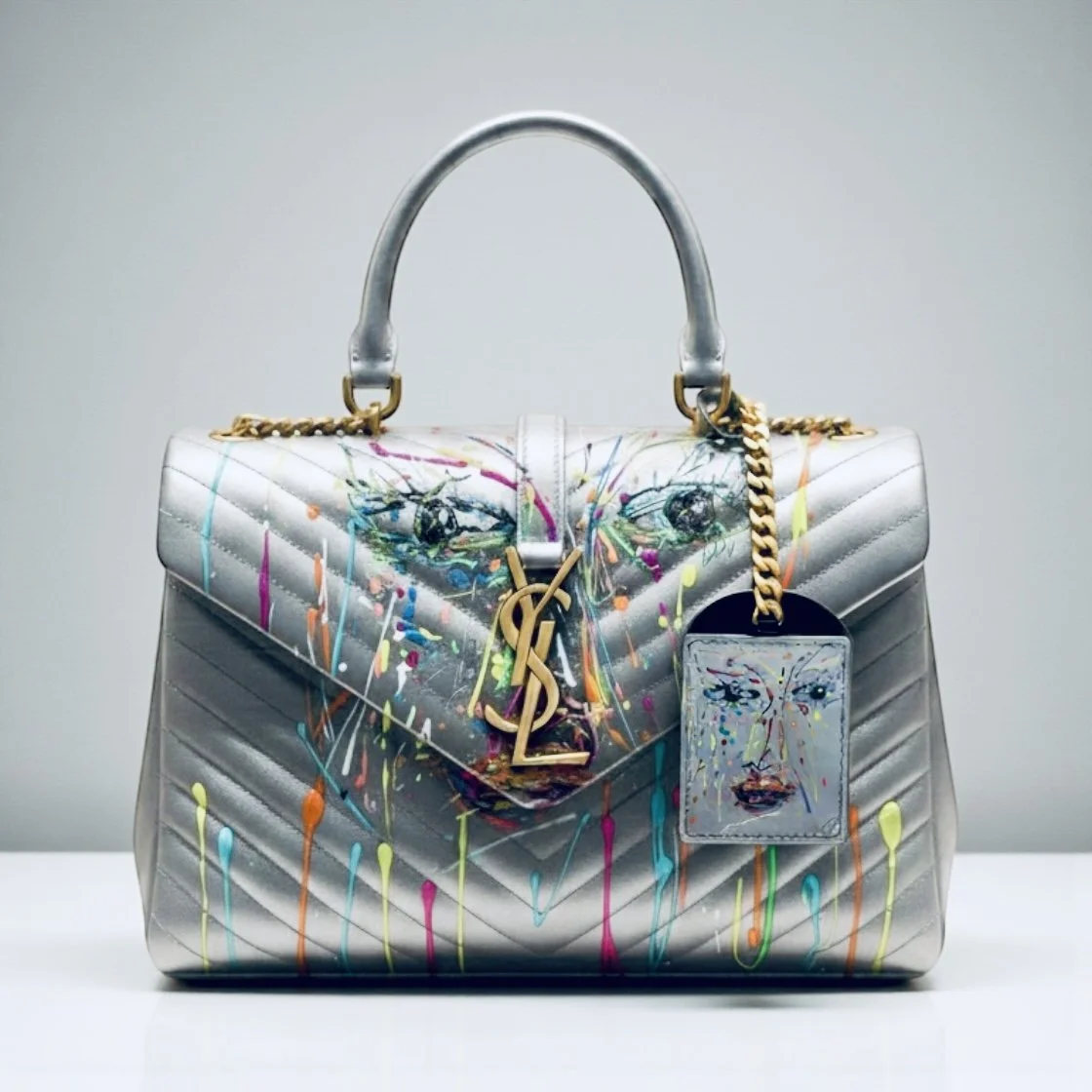 Borsa di design color argento con disegni pittorici colorati e una catena d'oro, con una grande targhetta con il volto dipinto, marchio Saint Laurent.