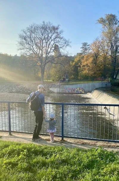 An man and young girl standing by a blue fence near a river, watching water flow over a small waterfall in a park with trees and graffiti in the background during late afternoon.