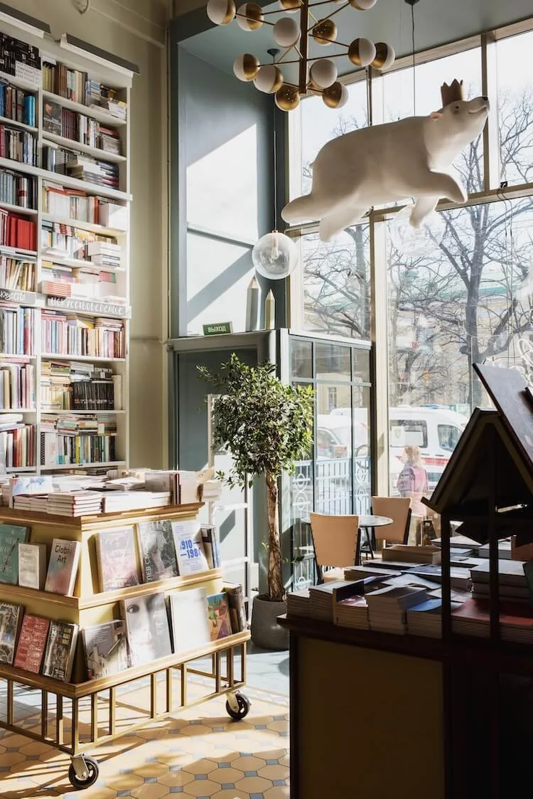 A narrow aisle in a cozy bookstore or library, lined with tall wooden shelves filled with books on both sides, a small wooden step stool in the middle, and a patterned rug on the floor. Warm lighting from ceiling bulbs illuminates the space.