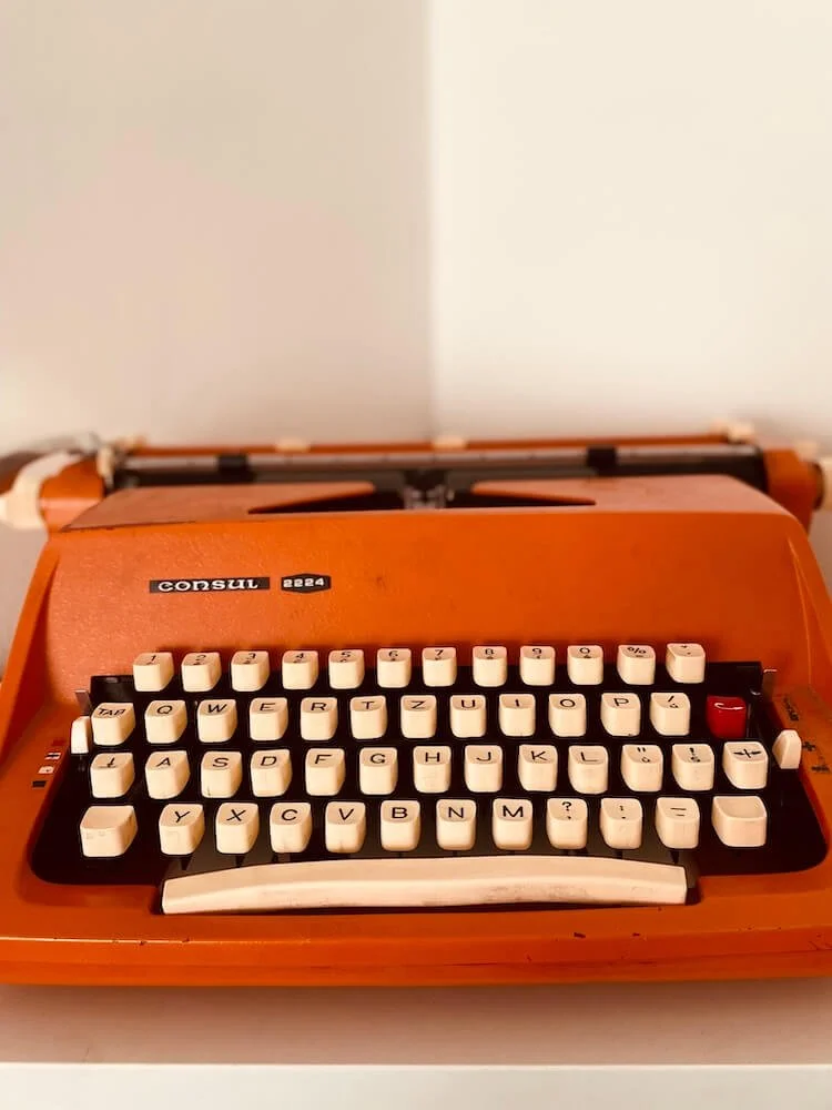 Orange vintage Consul 2224 typewriter with cream keys and a red key, placed on a white surface against a white wall.