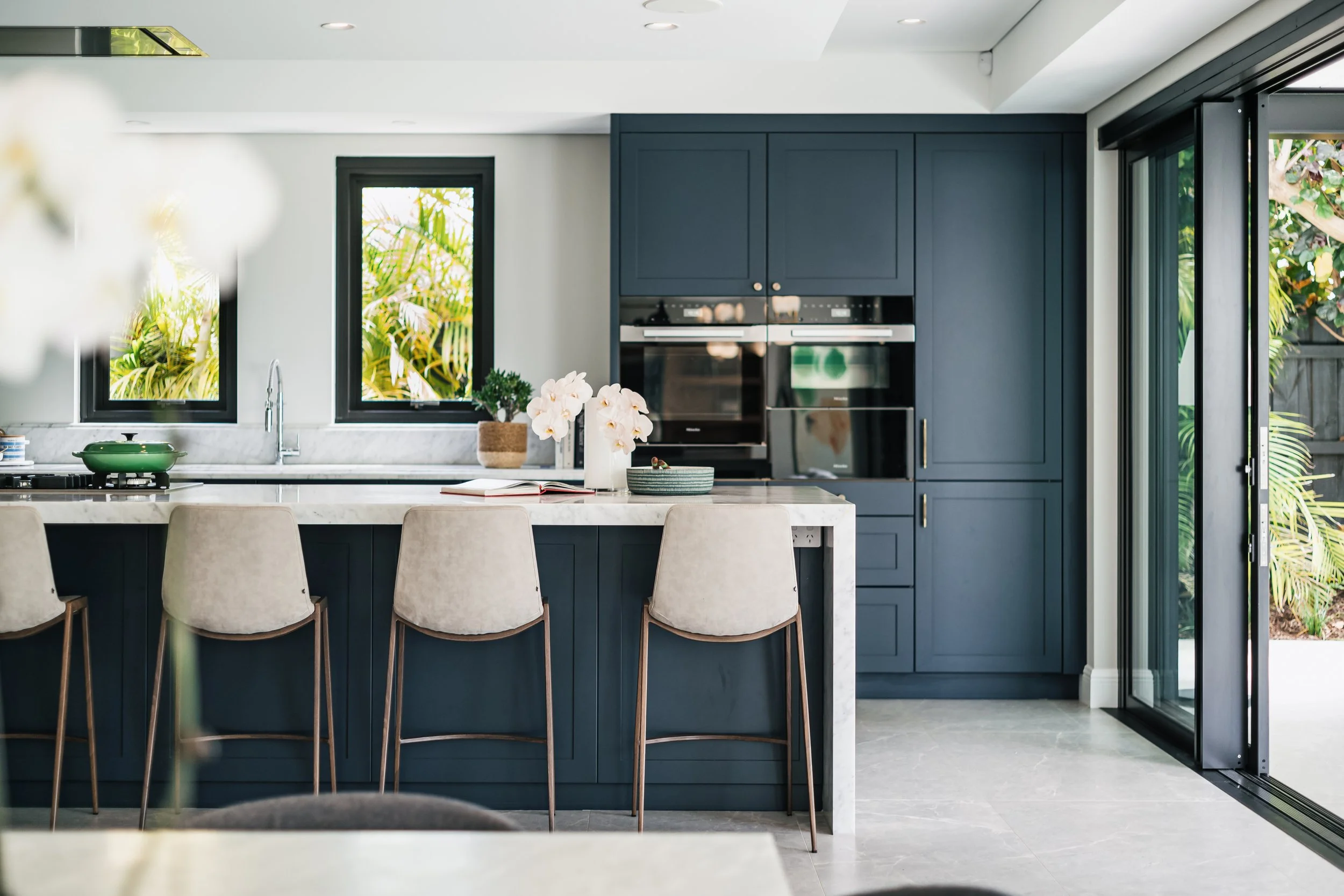 Modern kitchen with blue cabinets, white marble countertops, beige barstools, and large windows overlooking greenery, with a sliding glass door on the right.