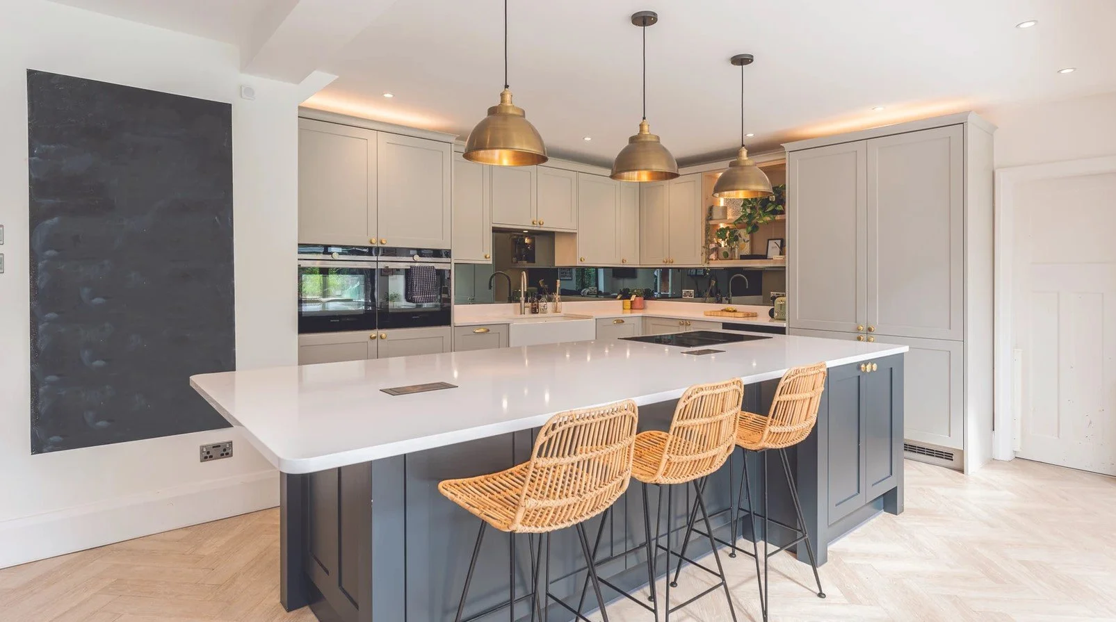 Modern kitchen with white countertops, light gray upper cabinets, dark gray lower cabinets, and three gold pendant lights. Wicker bar stools are at the island, and there are built-in appliances and greenery on shelves.