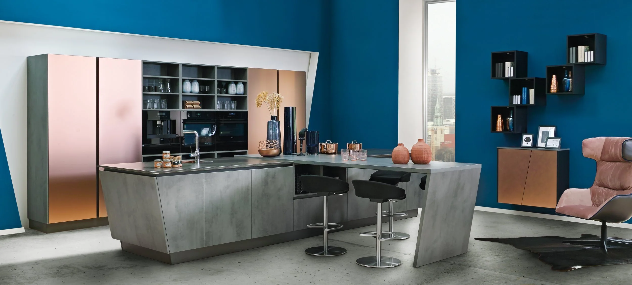 Modern kitchen with blue accent wall, gray kitchen island, pink and copper cabinets, black shelves with books and decor, and pink lounge chair with black rug.