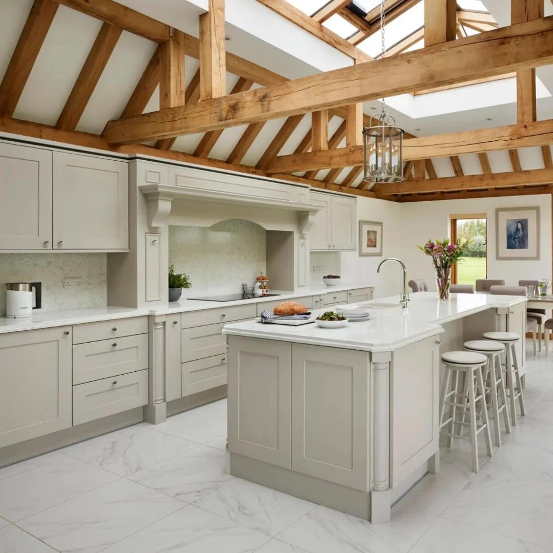 Modern kitchen with white cabinets, a marble kitchen island, wooden exposed beams, and a skylight ceiling. There are stools at the island, a vase with flowers, and a dining area in the background.