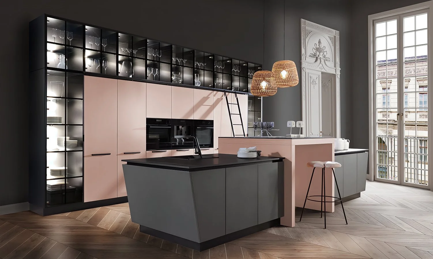 Modern kitchen with pink and gray cabinetry, open shelves with wine glasses and dishes, black countertops, wooden herringbone floor, large windows, and hanging woven light fixtures.
