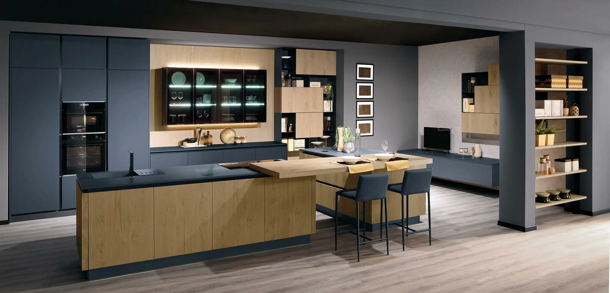 Modern kitchen with a combination of dark blue and light wood cabinets, a kitchen island with stools, built-in oven, glass display cabinet, open shelving with decorative items, small TV, and a wooden flooring.