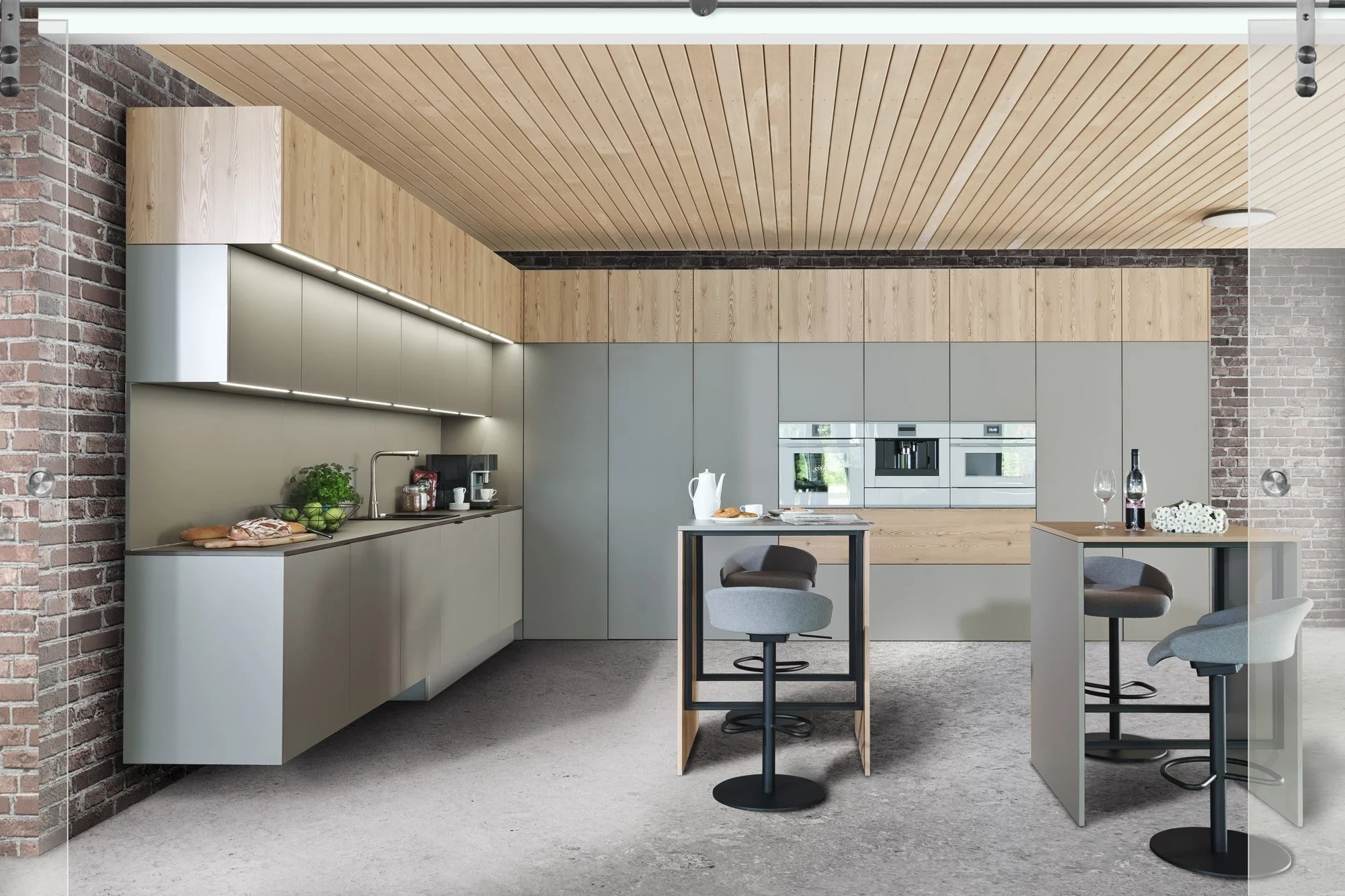Modern kitchen with beige cabinetry, wooden ceiling, brick wall, and gray barstools at small high tables. Counter with food and drink items.