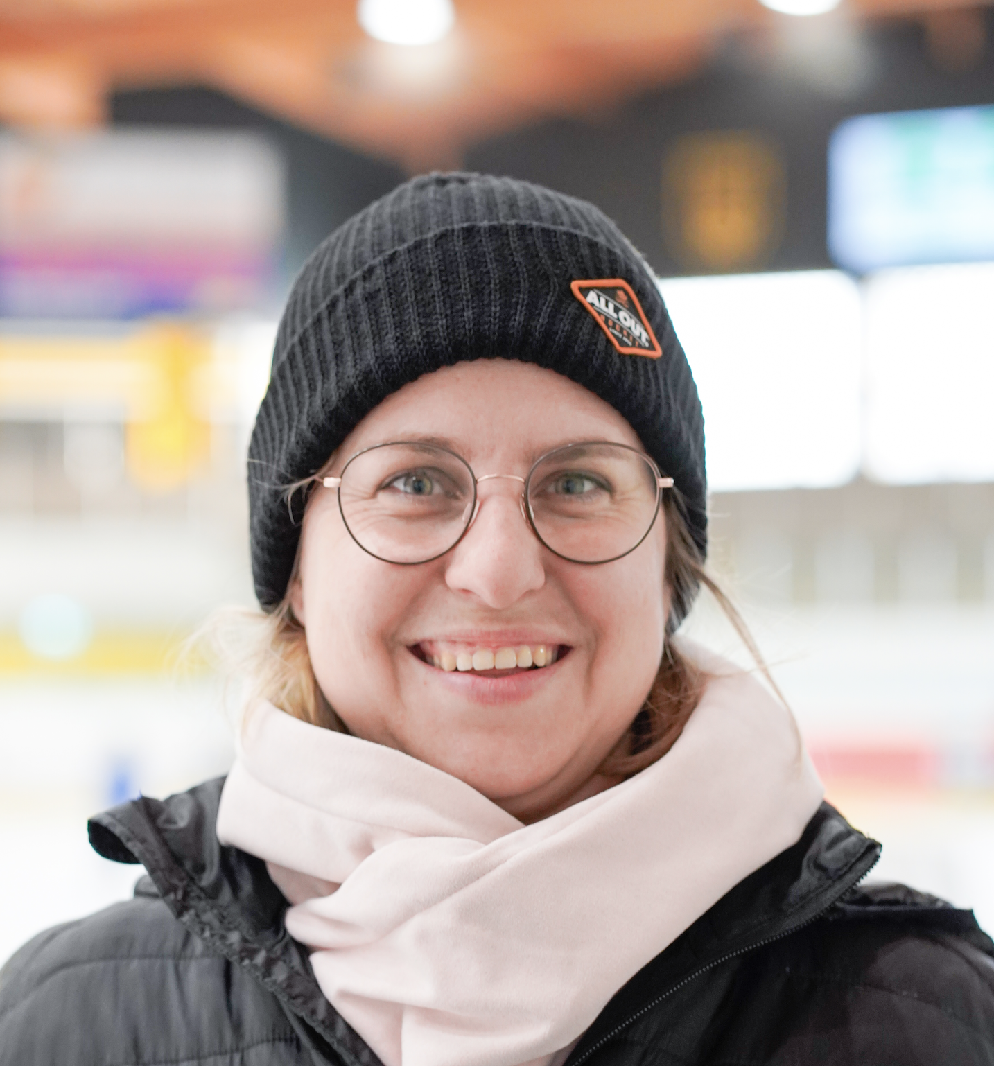 Ein lachender junger Mann mit Brille, schwarzer Mütze und Winterjacke im Eislaufstadion.