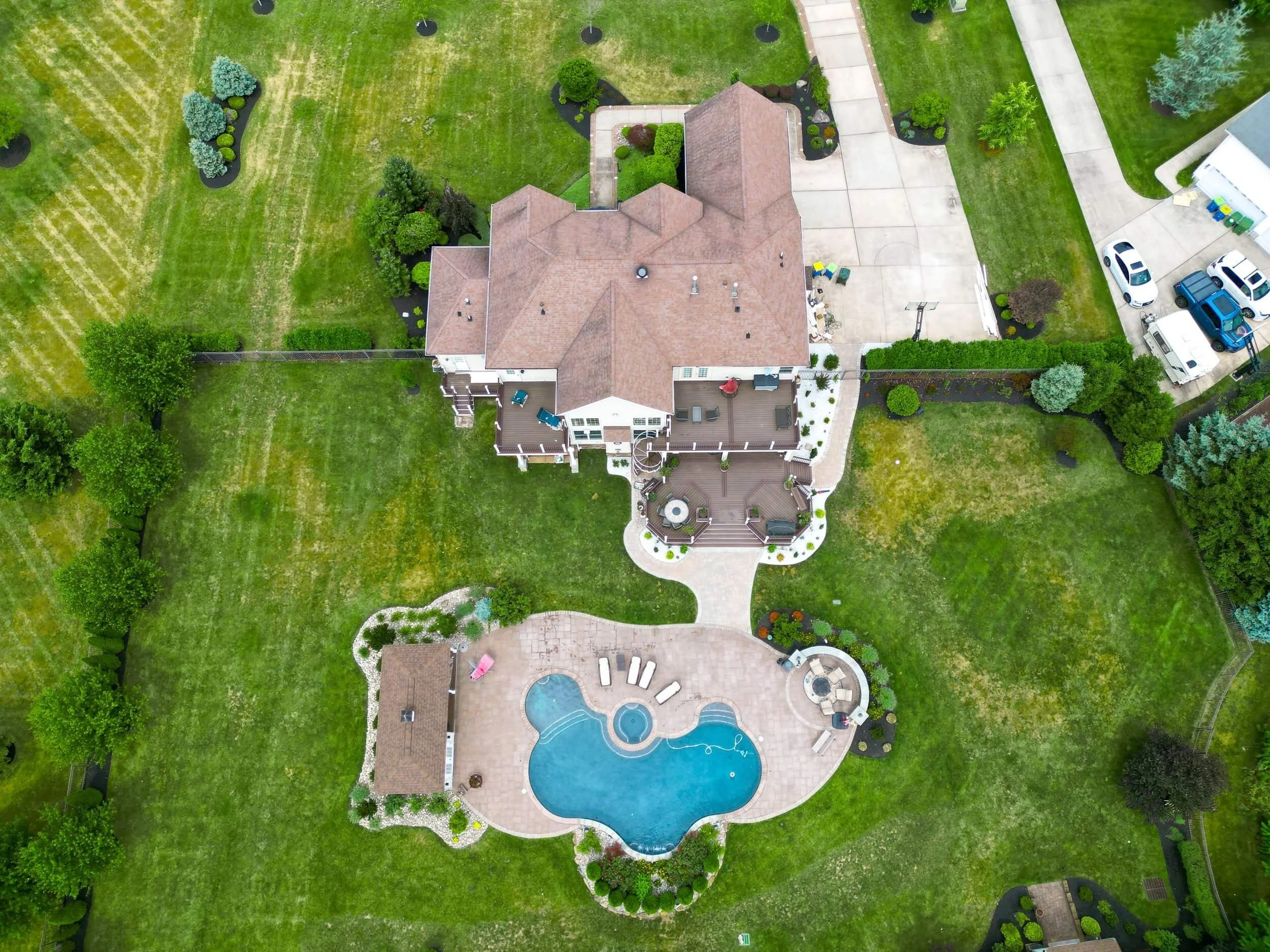 Aerial view of a large house with a backyard featuring a swimming pool, patio, and lawn. The house has a brown roof, multiple decks, and is surrounded by landscaped greenery. There are several driveways and parked cars.