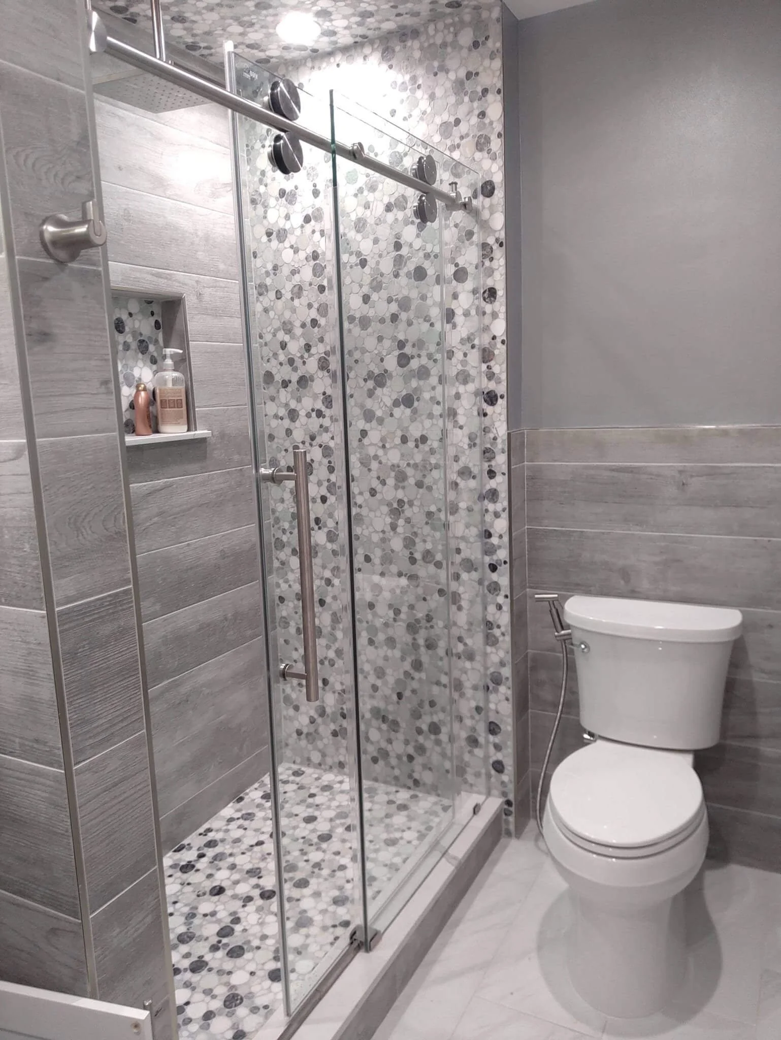 Modern bathroom with a glass-enclosed shower and toilet. The shower features gray and white mosaic tiles, a sliding door, and built-in shelving. The walls are finished with gray tiles, and the bathroom has bright lighting.