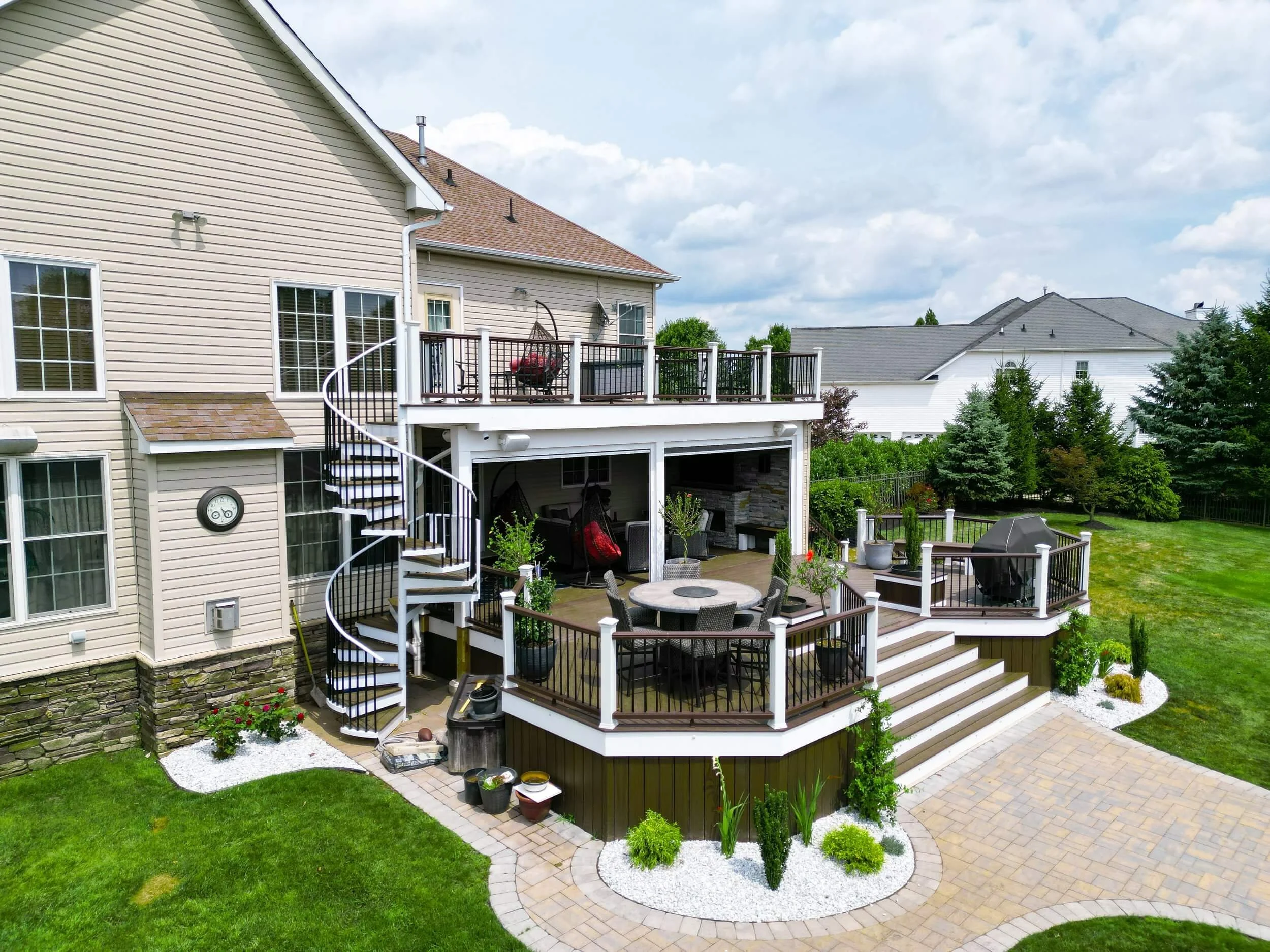 Aerial view of a wooden deck with outdoor furniture, plants, and a grill. The deck features a round table with chairs and multiple planters. A spiral staircase is visible on the left, and a stone walkway surrounds the deck with white rock landscaping.
