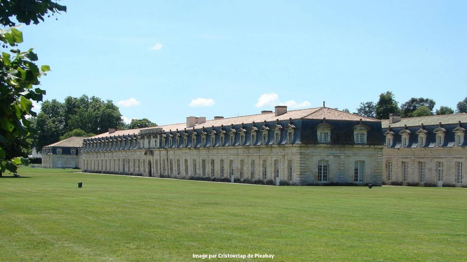 Un grand bâtiment classique en pierre avec un toit en ardoise et de nombreuses fenêtres, entouré d'une grande pelouse verte et d'arbres, en plein jour avec un ciel bleu clair.