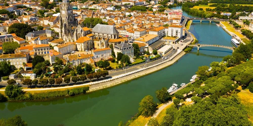 Vue aérienne d'une ville avec une rivière traversant la centre-ville, entourée de bâtiments en pierre, d'églises avec des toits en ardoise, et des espaces verts. Saintes