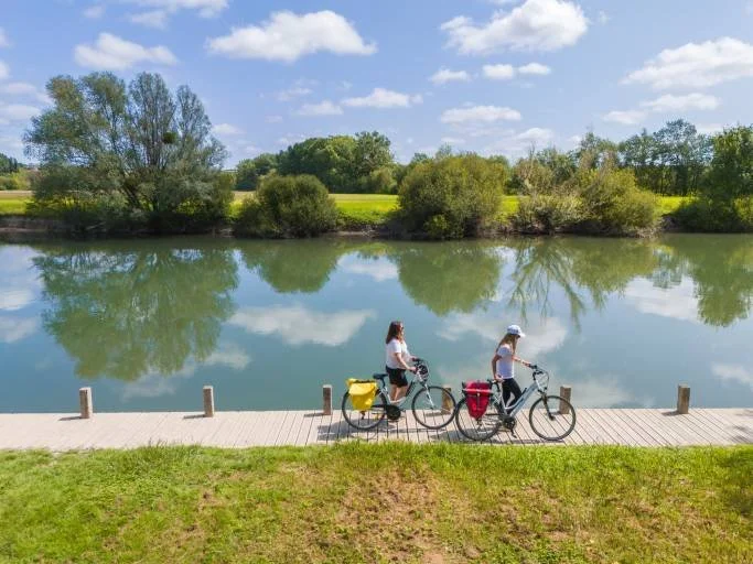 Deux personnes faisant du vélo le long d'une rivière sous un ciel bleu avec des nuages blancs, entourées d'arbres verts et d'un paysage naturel.