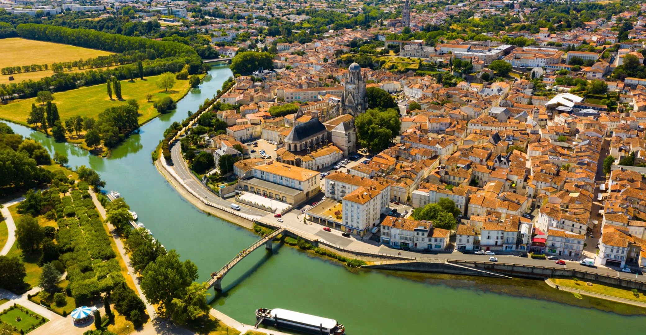 Vue aérienne d'une ville avec une rivière serpentant à travers le centre, bâtiments aux toits en tuiles orange, espaces verts et un pont sur la rivière. Saintes