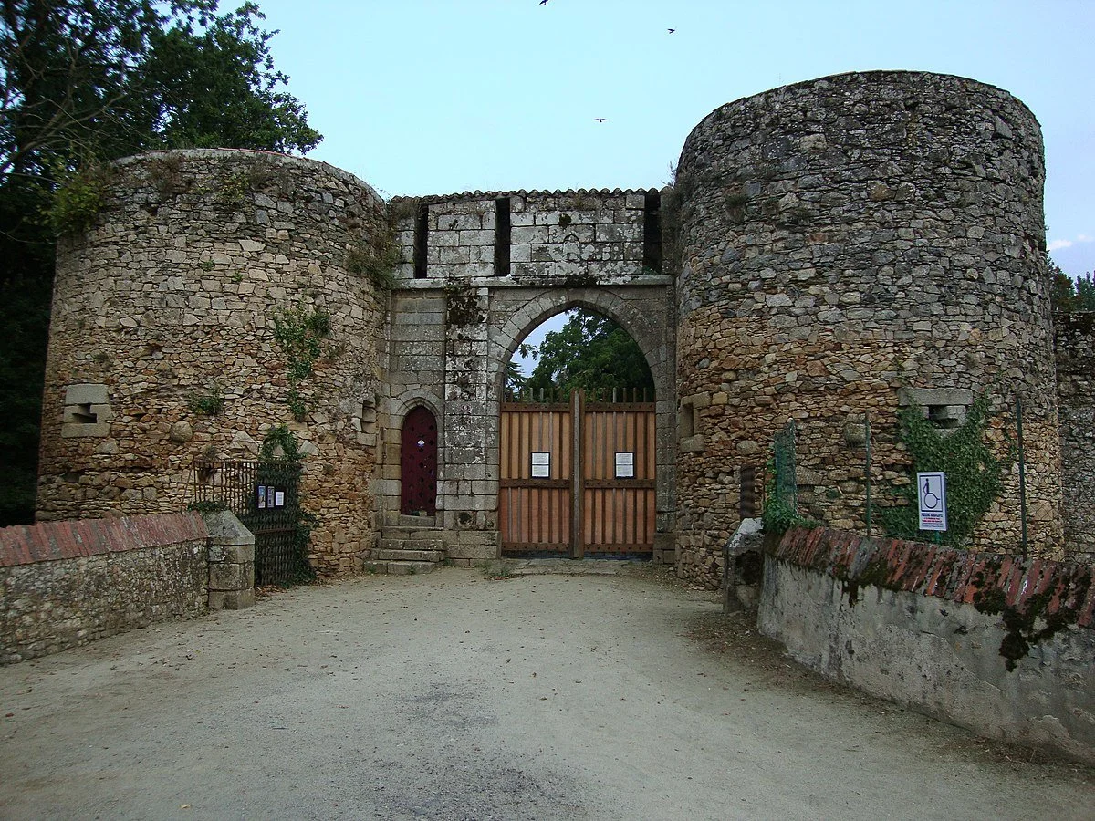 Old fortified stone gate with two round towers, a wooden door and an archway. Earth path in front. Accessibility and restriction signs.